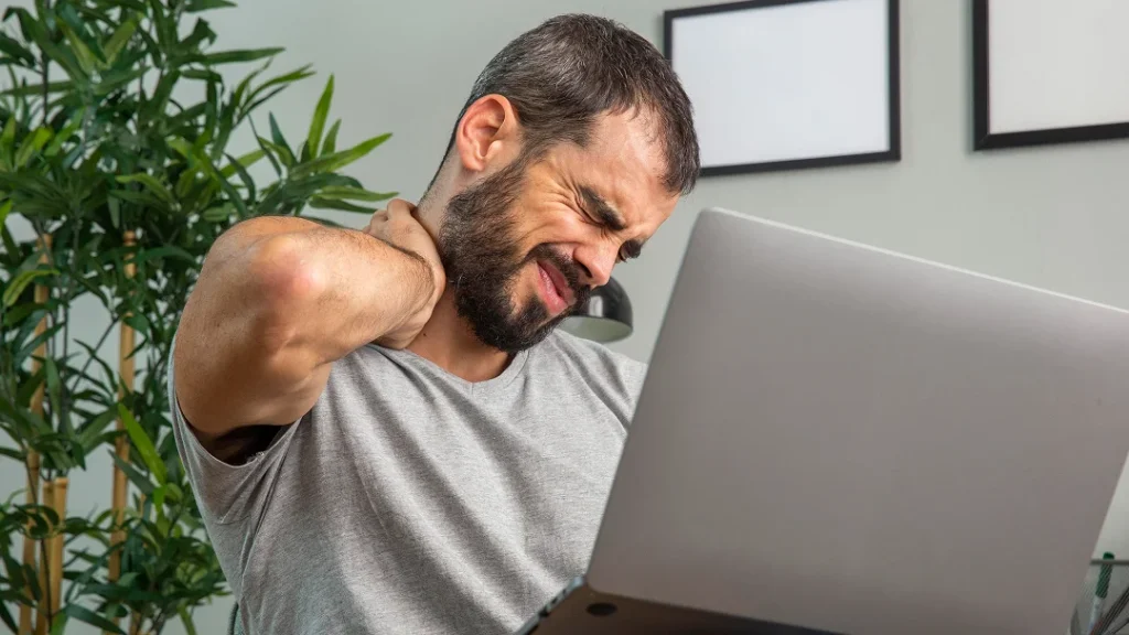 Un homme assis devant un ordinateur portable, se tenant le cou avec une expression de douleur.