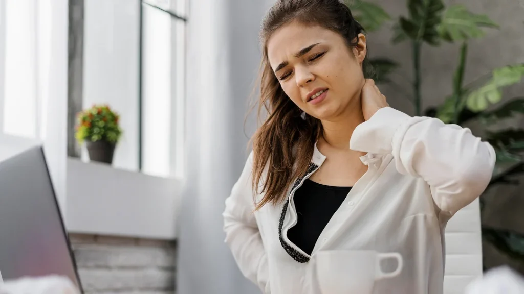 Une femme souffrant de douleurs cervicales assise à son bureau, visiblement inconfortable.